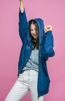 一位年轻女子面带微笑，身着蓝色连帽衫，戴着耳机，在粉色背景下随音乐起舞，尽显风格