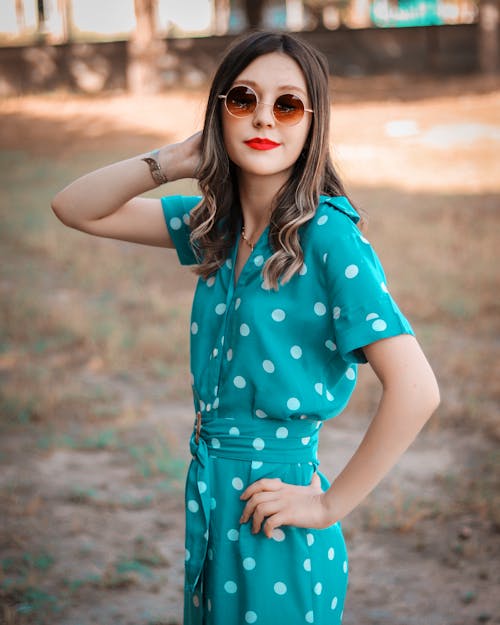 Side view of positive young female in trendy dress and sunglasses touching hair while resting in countryside on sunny day