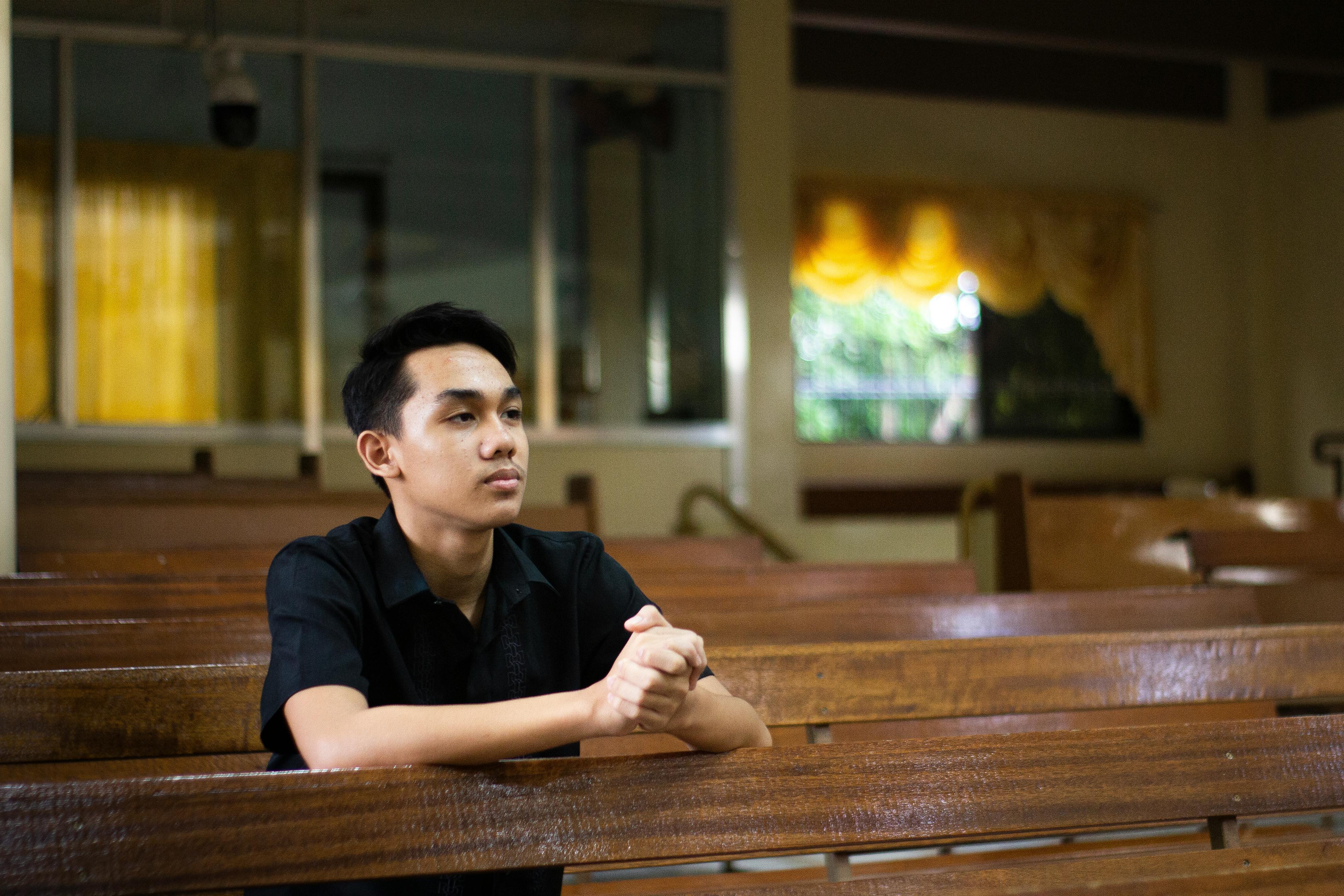 一个年轻人坐在教堂的长凳上，双手紧握祈祷