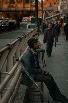 一位穿着休闲服、拄着拐杖坐在街边长凳上抽烟的中年少数族裔男性侧面图，他正在深思熟虑