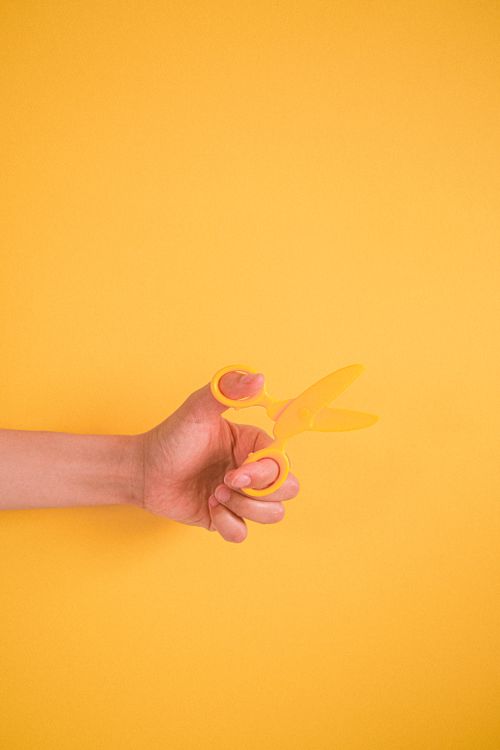 Free Minimalist image of a hand holding yellow scissors on a yellow background. Stock Photo