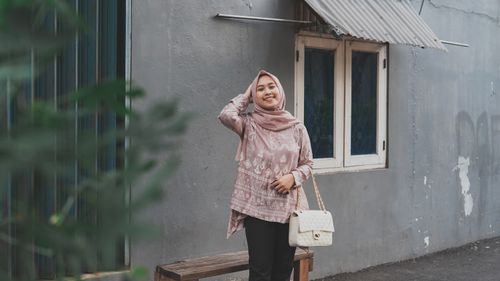 Free A happy woman in a pink hijab and stylish outfit standing against a grey urban building. Stock Photo