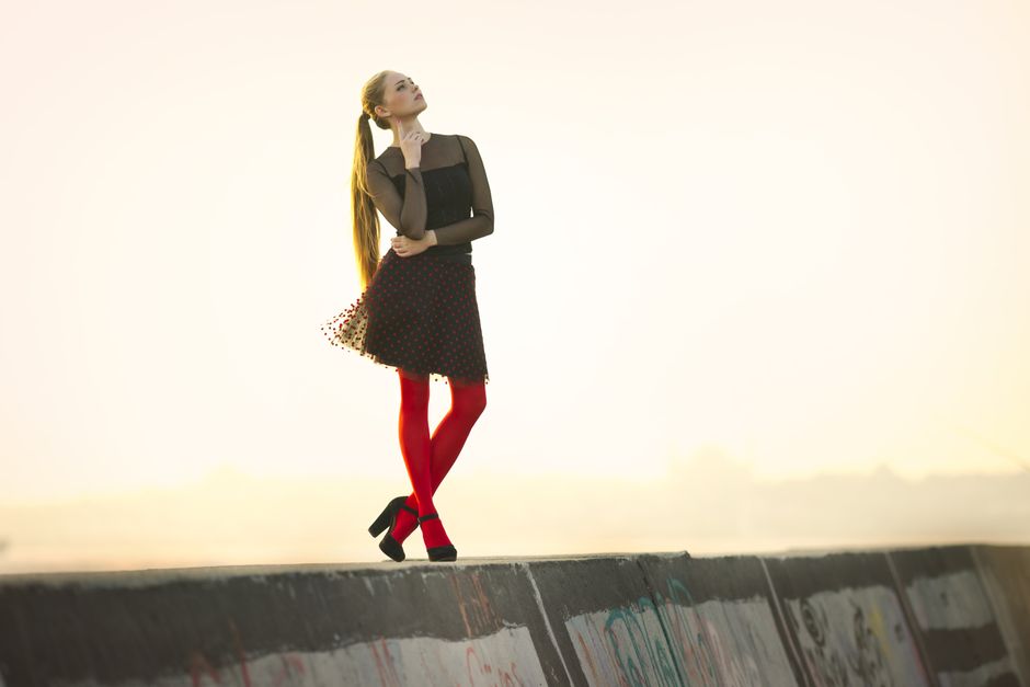 A fashionable woman in a black dress and red tights poses on a concrete barrier outdoors during the day.