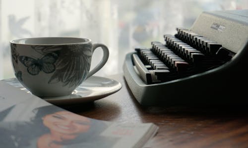 White and Blue Ceramic Cup beside Typewriter on Brown Wooden Table