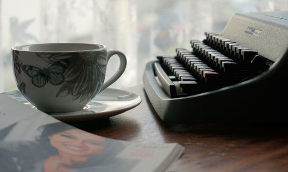 A cozy still life featuring a vintage typewriter and floral coffee cup by a window.