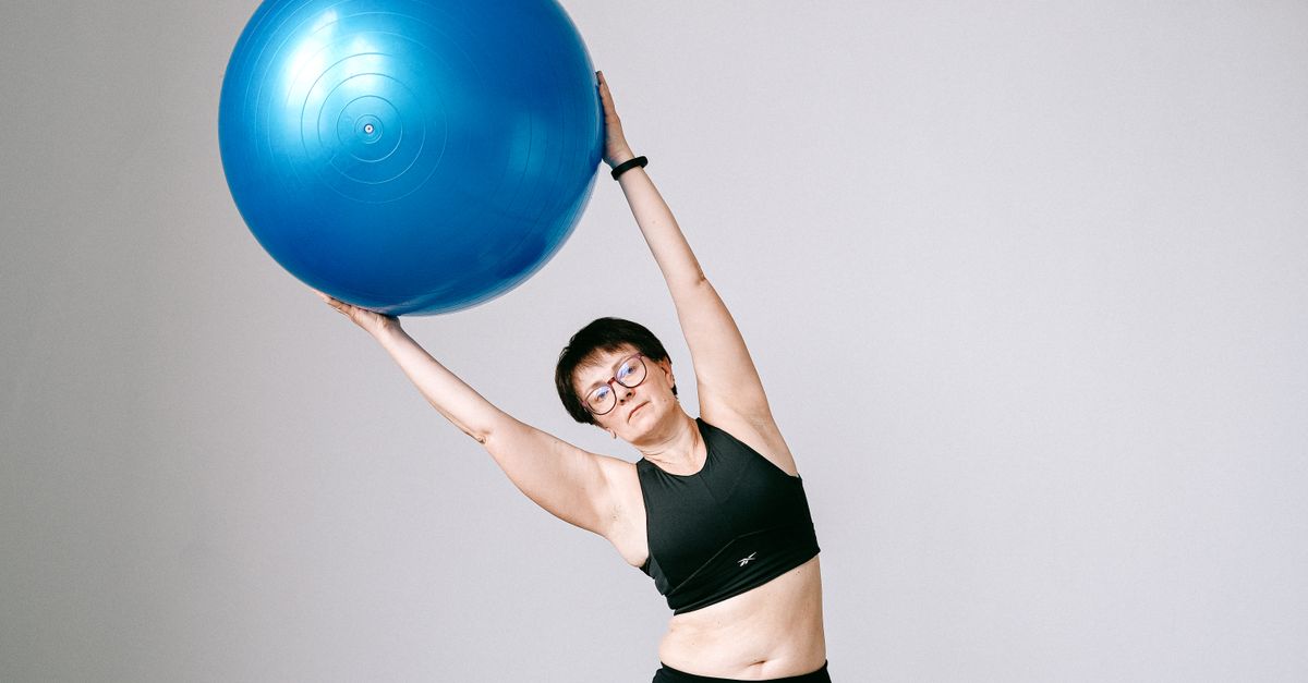 Adult woman in sportswear exercising with blue stability ball against gray background.