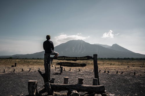 Man Sitting on the Baluran National Park Sign and looking at a Mountain 