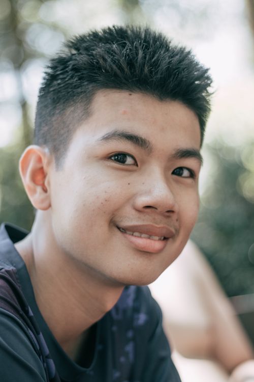 Free Cheerful Asian teenage boy in dark shirt with short black hair looking at camera with friendly toothy smile in forest on blurred background Stock Photo