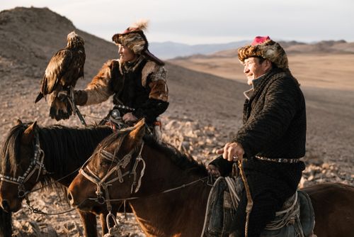 Free Two eagle hunters in traditional attire riding horses in a desert landscape, showcasing ancient hunting traditions. Stock Photo