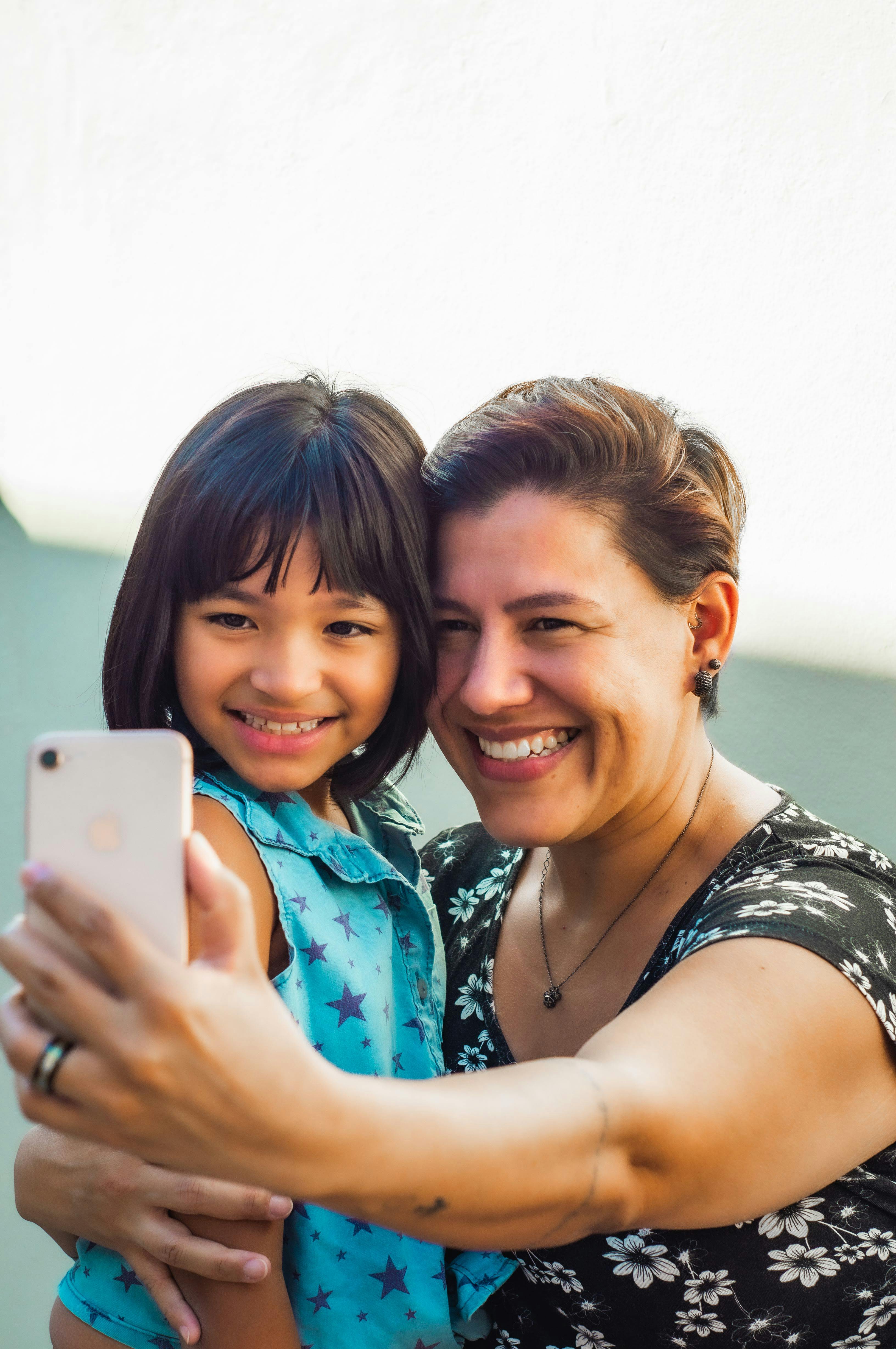Happy middle aged woman hugging adorable Asian girl and taking selfie on mobile phone together