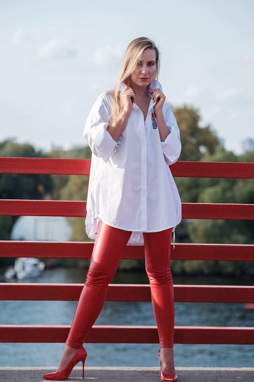 Free Full body of young confident female in high heeled shoes and white shirt standing on bridge over river and looking at camera Stock Photo
