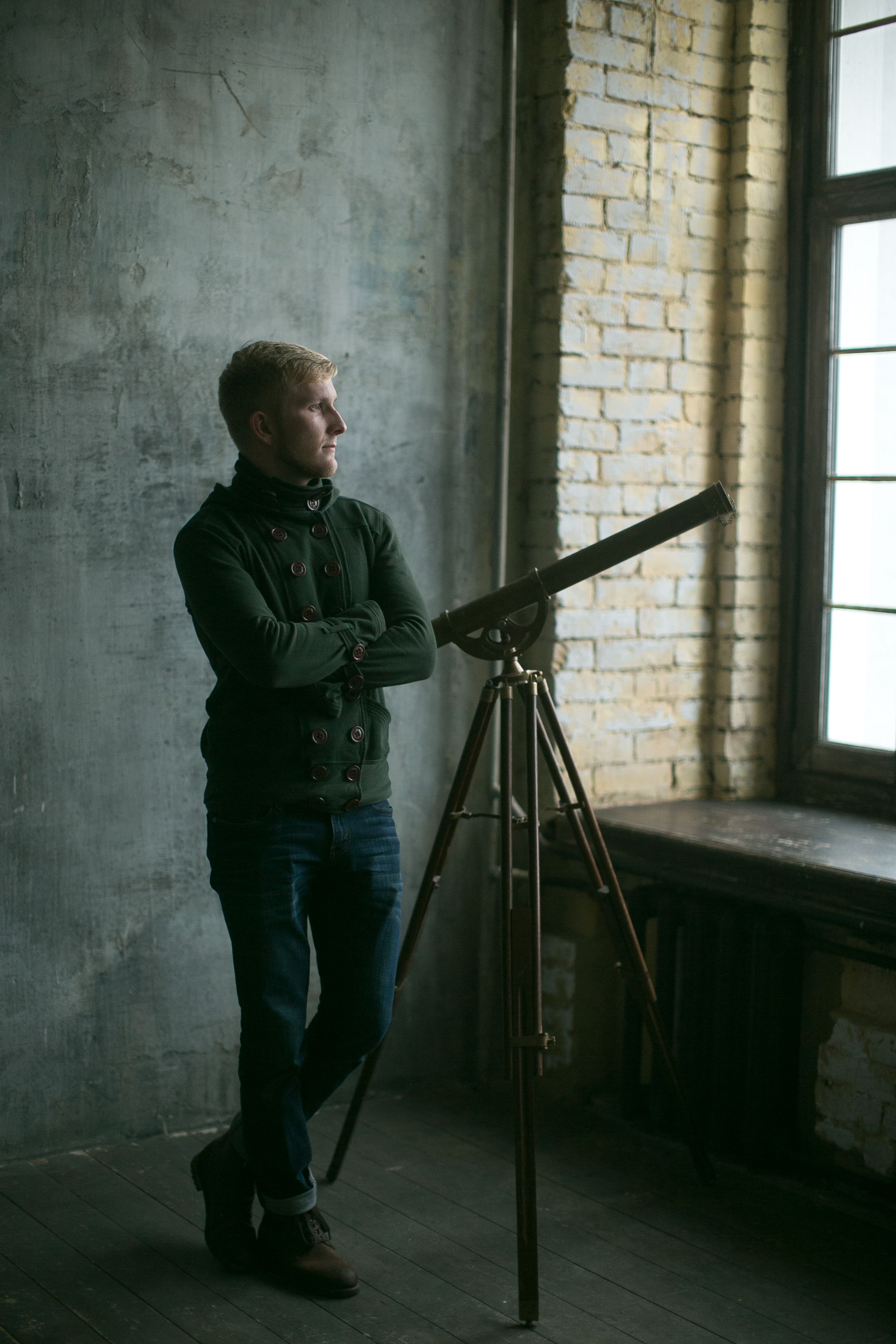 Man standing indoors beside vintage telescope and window with industrial decor.