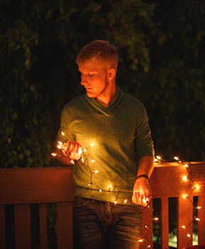 A man in a green sweater holds glowing string lights outdoors at night.
