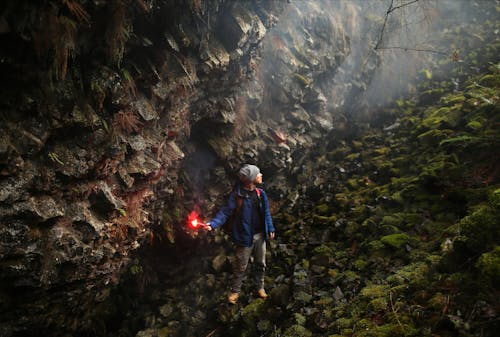 Free A Man in Blue Jacket Holding a Flare While Standing on the Mossy Rocks in the Cave Stock Photo