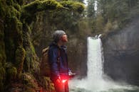 A Man Wearing Blue Jacket Standing Near the Waterfall While Holding a Flare