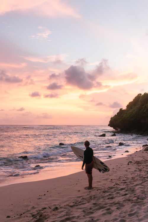 Free A lone surfer standing on a sandy beach, gazing at the colorful sunset sky. Stock Photo