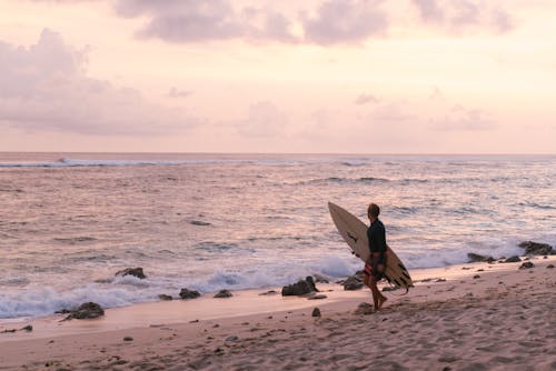 Free Man in Black Shirt Carrying White Surfboard Walking on Beach Stock Photo