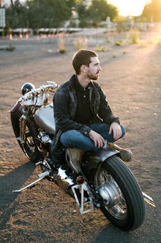 A young man with a beard sits casually on a vintage motorcycle during a golden sunset.