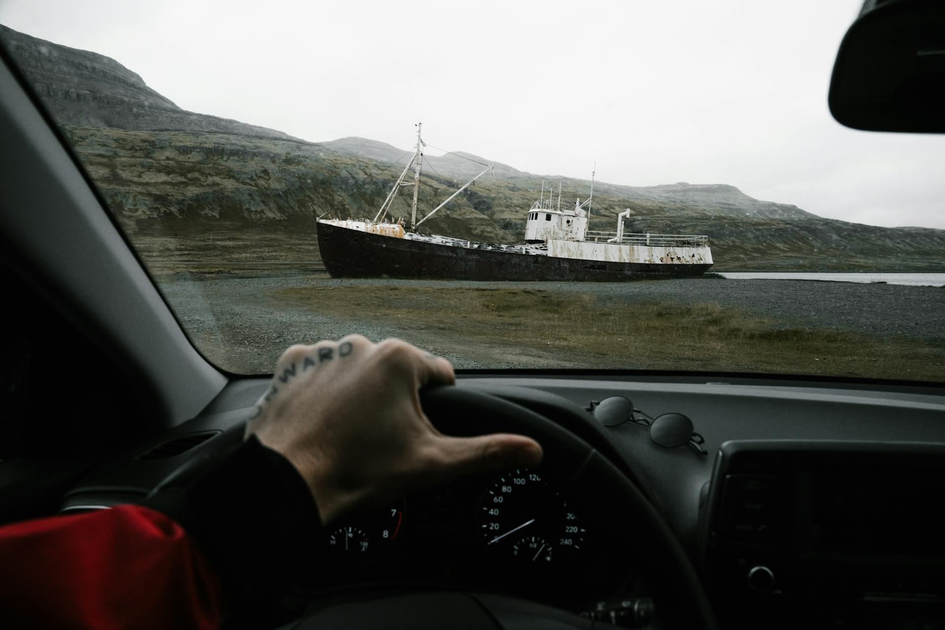 Driver views an abandoned, rusty shipwreck on a rugged coastline through car window, weather overcast.