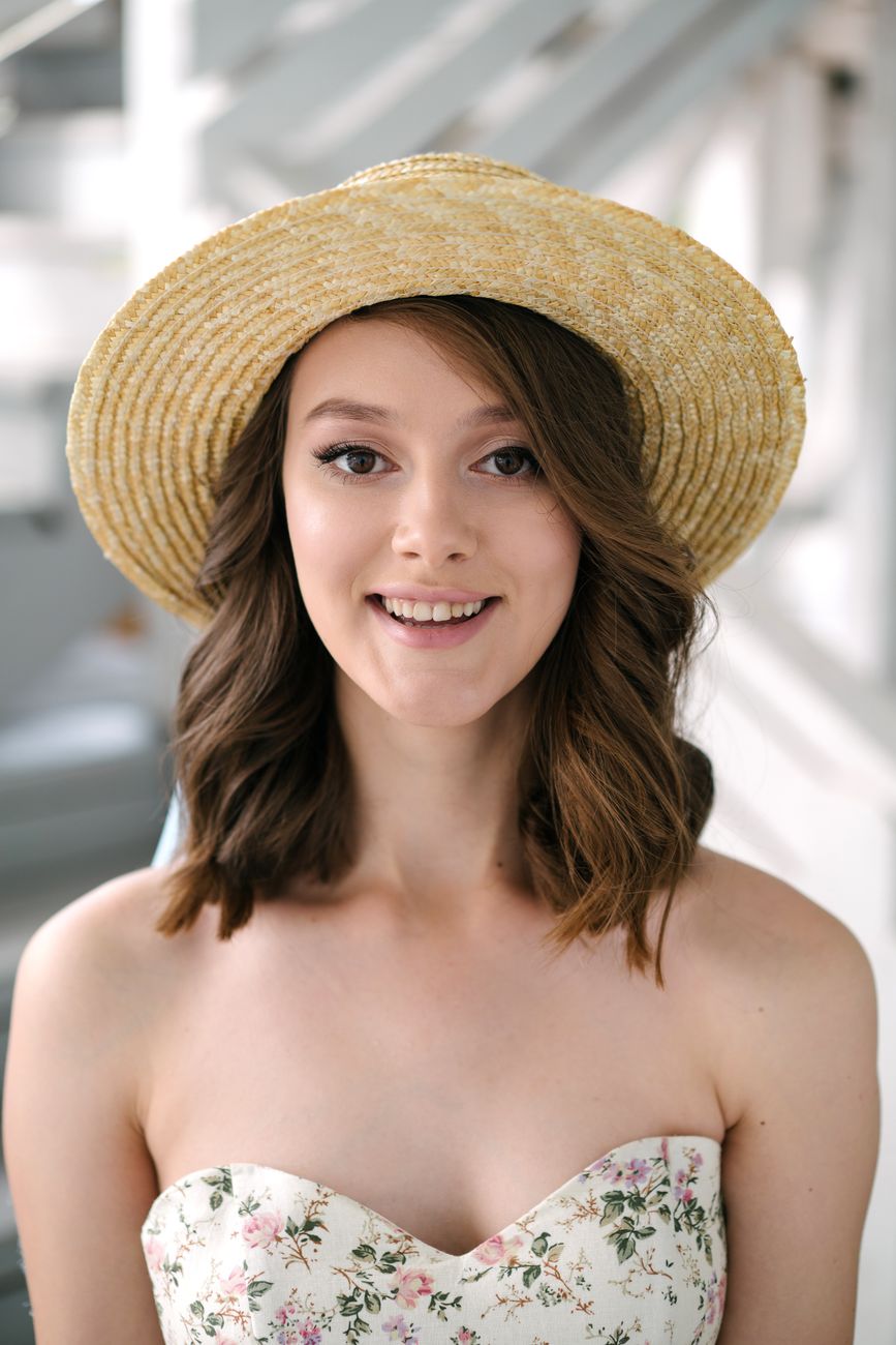 Charming young woman wearing a straw hat and floral dress, smiling warmly.