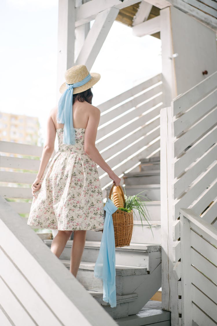 Woman In A Floral Dress And Straw Hat Carrying A Basket And Walking Up The Stairs 