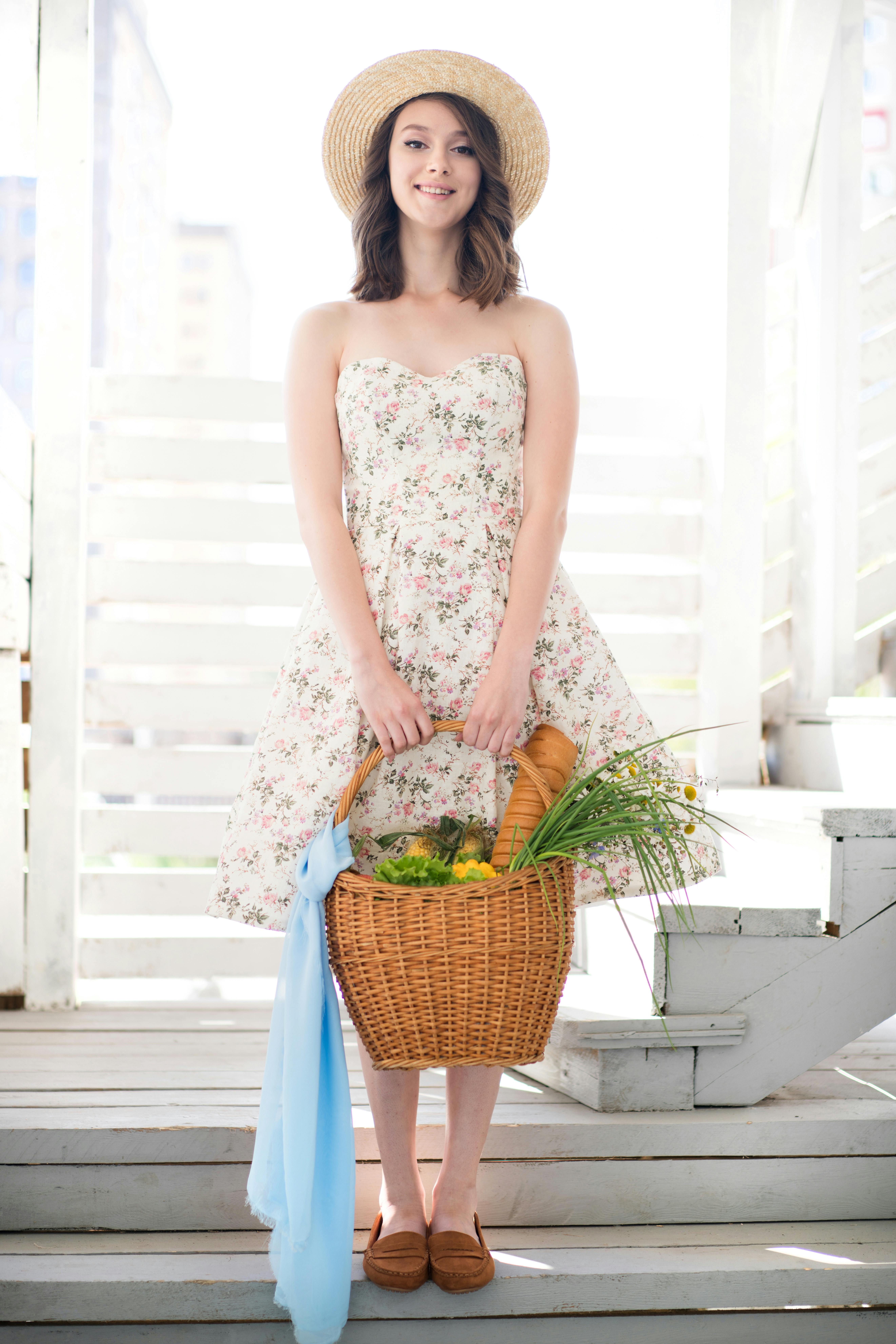 Woman in floral dress with basket, wearing a straw hat in a bright, summer setting.