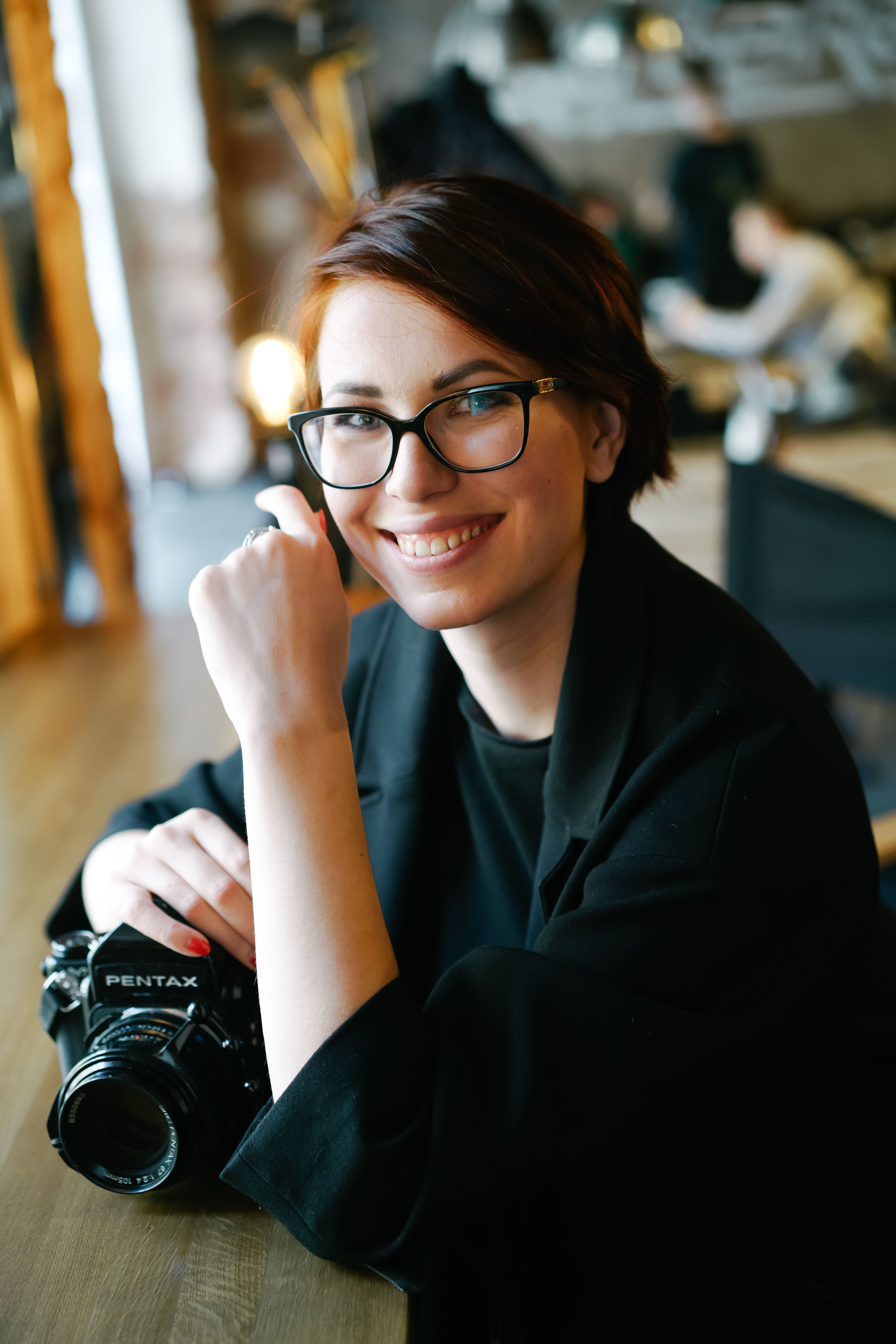 Portrait of a smiling woman wearing eyeglasses, holding a camera in a cozy indoor cafe environment.