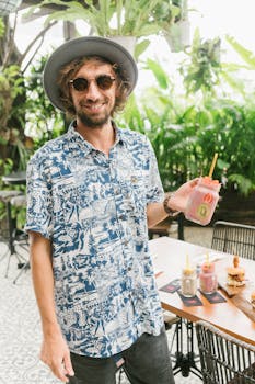 Casually dressed man in sunglasses and hat holding a fresh fruit drink outdoors.