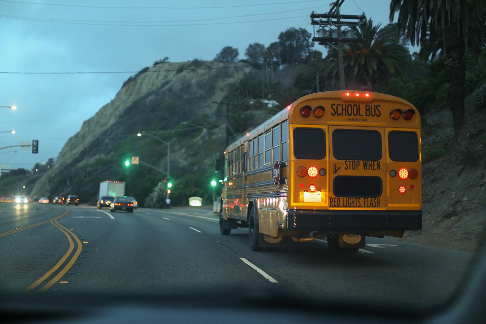 Yellow school bus travels along a scenic highway near cliffs at dusk, showcasing everyday transportation.