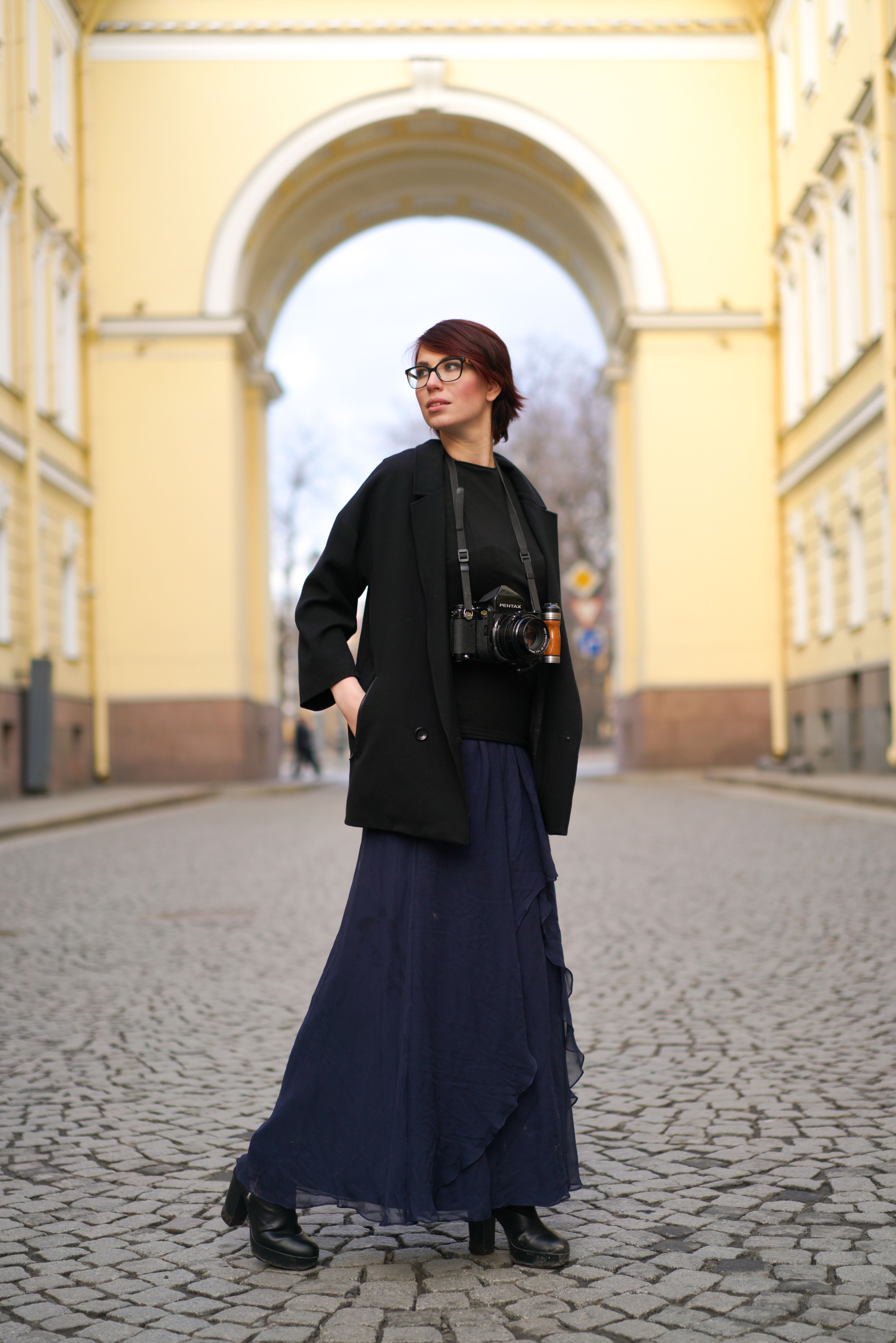 Woman with camera stands confidently under a yellow arch on a cobblestone street.