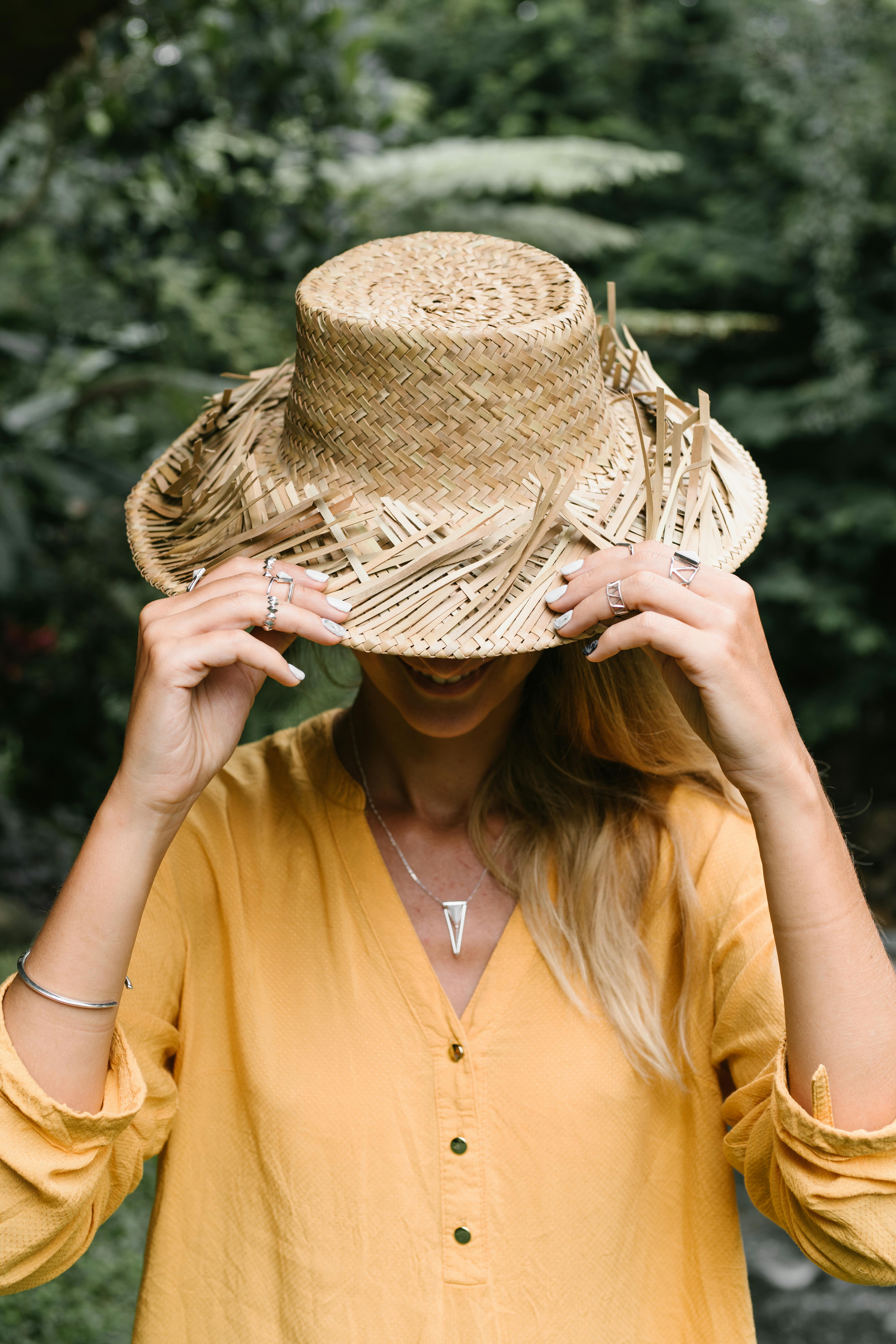 Playful woman conceals her face with a straw hat in a lush green outdoor setting.