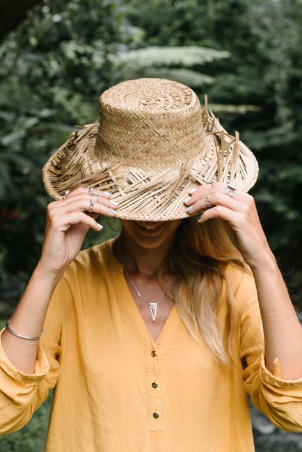 Playful woman conceals her face with a straw hat in a lush green outdoor setting.
