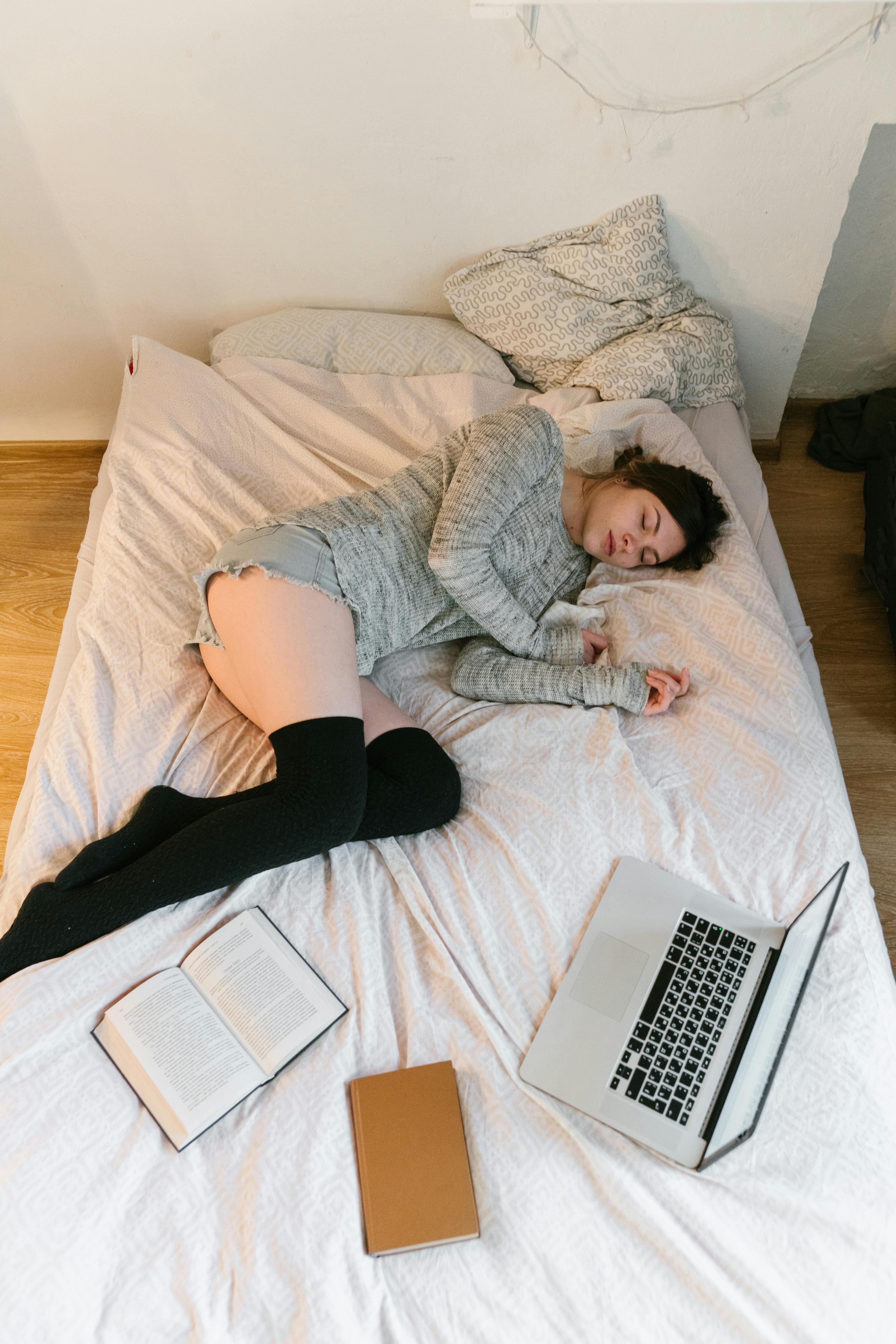 女人躺在舒适房间的床上睡觉，手里拿着打开的笔记本电脑和书籍