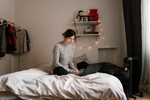 Woman in Gray Sweater Sitting on Bed Using Macbook