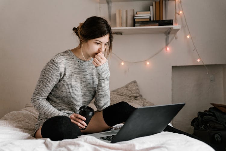 Confused Woman In Gray Sweater Sitting On Bed Using Macbook