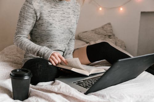 Woman in Gray Sweater and Black High Socks Sitting on Bed with a Book and Laptop