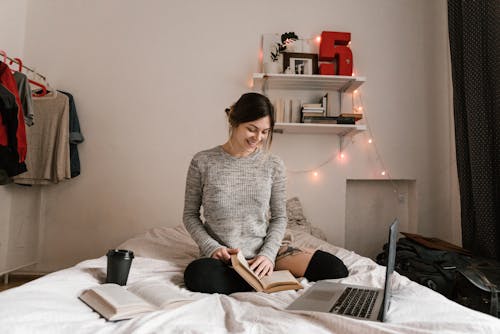 Smiling Woman in Gray Sweater Sitting on Bed with Books and Laptop