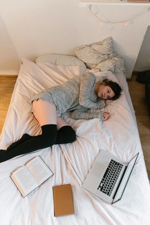 Free Woman in Gray Sweater Lying on Bed with Books and a Laptop Stock Photo