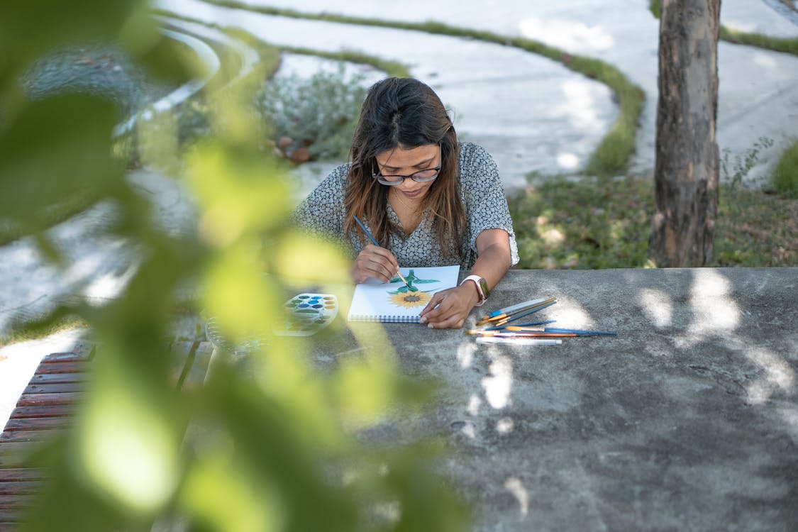 Focused young woman painting flower in notebook sitting in garden