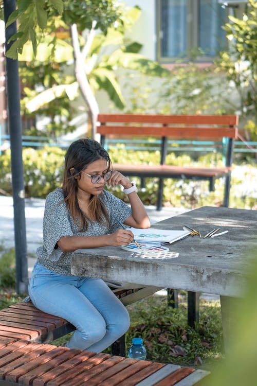 Serious young casual ethnic female painter in eyeglasses sitting on bench in park and drawing with watercolors and brushes