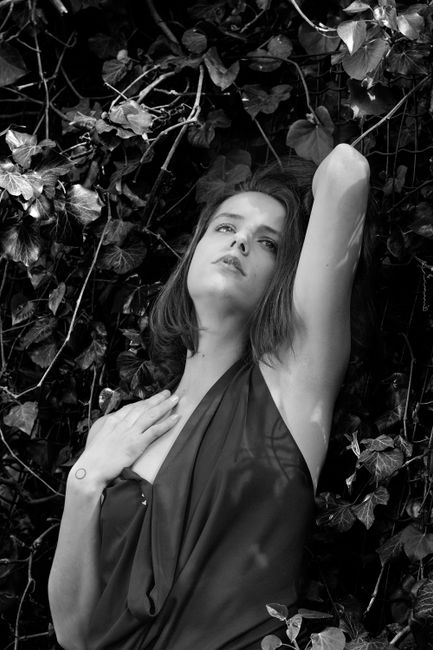 Black and white of young sensual woman in light top standing near plants in garden with hand behind head and looking away thoughtfully