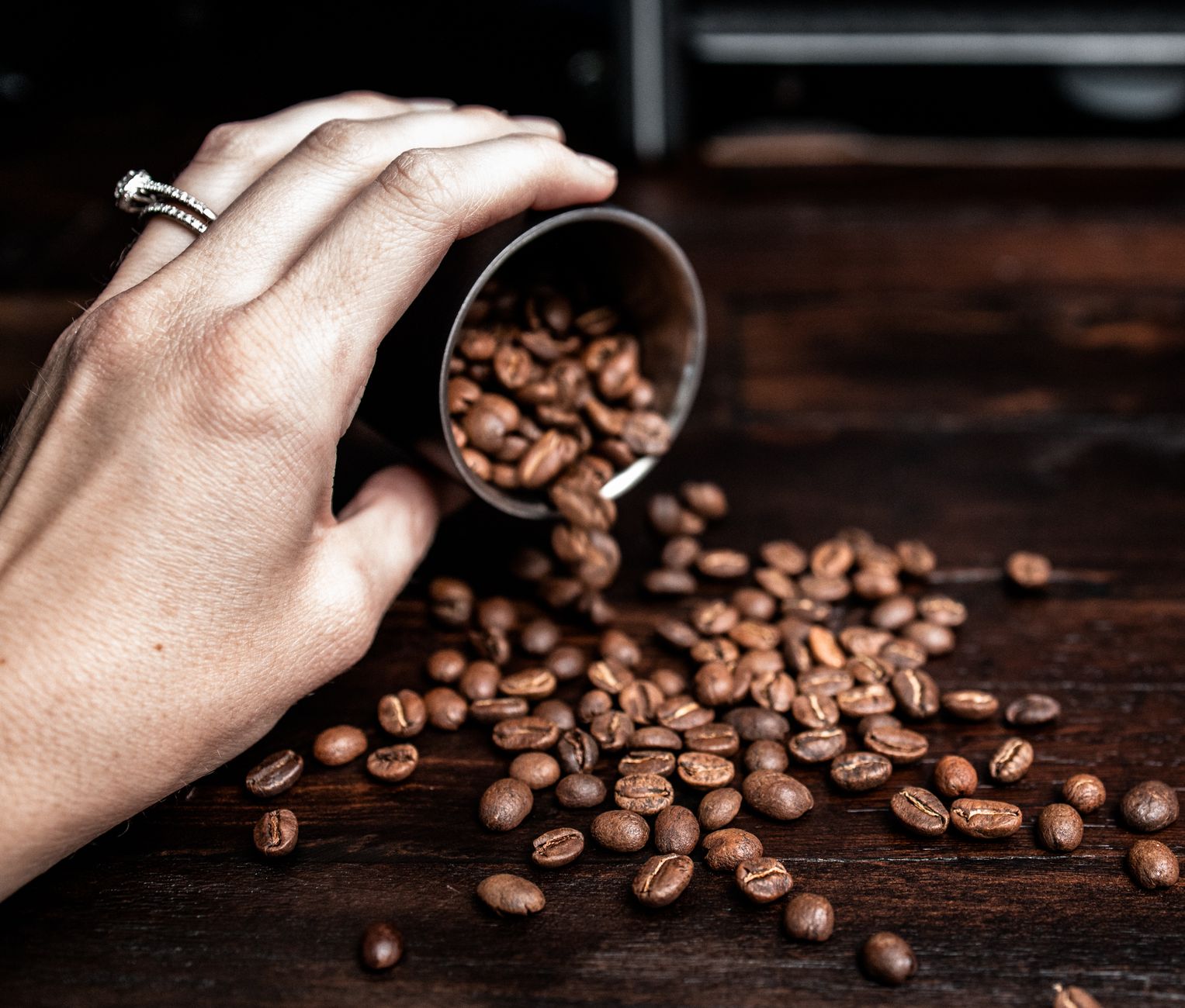 Crop anonymous female holding metal cup and pouring coffee beans on wooden table