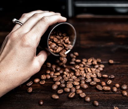 A hand pours aromatic coffee beans from a container onto a wooden surface, highlighting rich flavor and aroma.