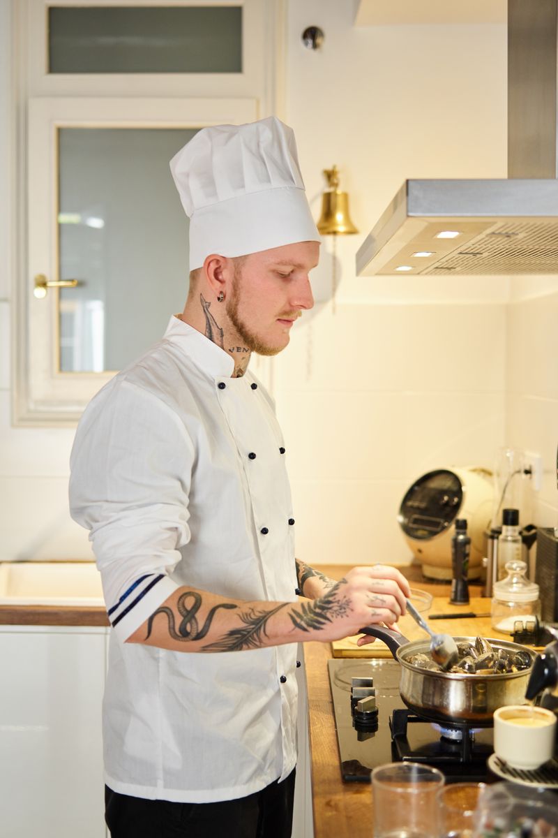 Tattooed chef in a white uniform preparing food in a sleek kitchen.