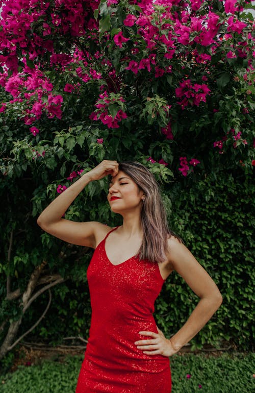 Smiling young female wearing red dress with bare shoulders standing with closed eyes and hands on waist and head in garden