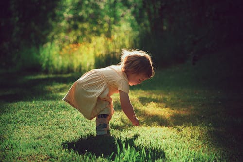 Side view of adorable kid in dress leaning forward on bright green meadow with shade while looking down in back lit