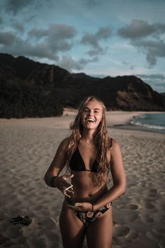 一名女子身着泳衣，在风景优美的海滩上微笑，那里有郁郁葱葱的山脉和多云天空