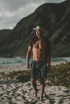 Shirtless man in a hat walking on a sandy beach by the sea with mountains in the background.
