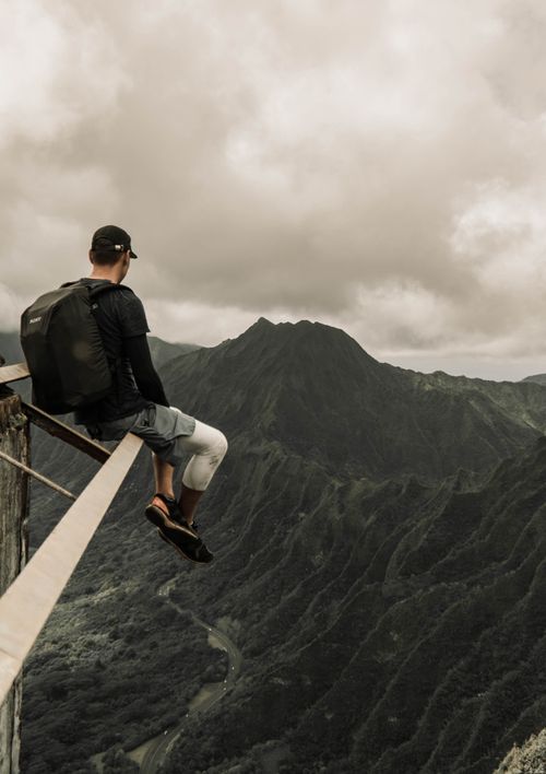 Free A man sits daringly on the edge of a mountain, enjoying breathtaking views of a lush valley. Stock Photo