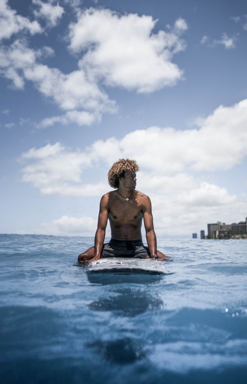 Free A young man sitting on a surfboard in the ocean under a vibrant blue sky with clouds. Stock Photo